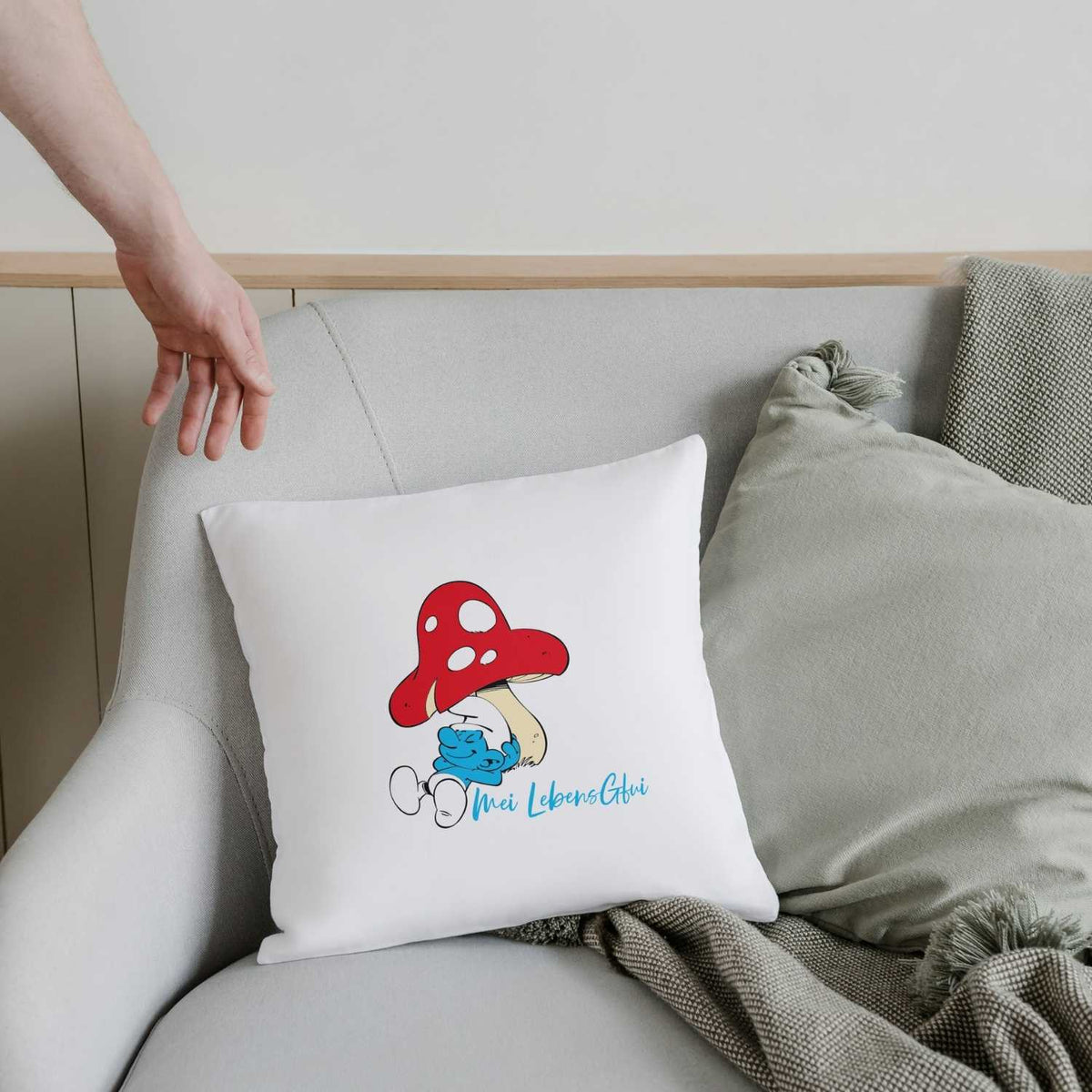 Weißes ®Die Schlümpfe "Do legst di nieda!" Kuschelkissen mit Motiv des liegenden Schlumpfs unter einem Fliegenpilz auf einem Sofa.