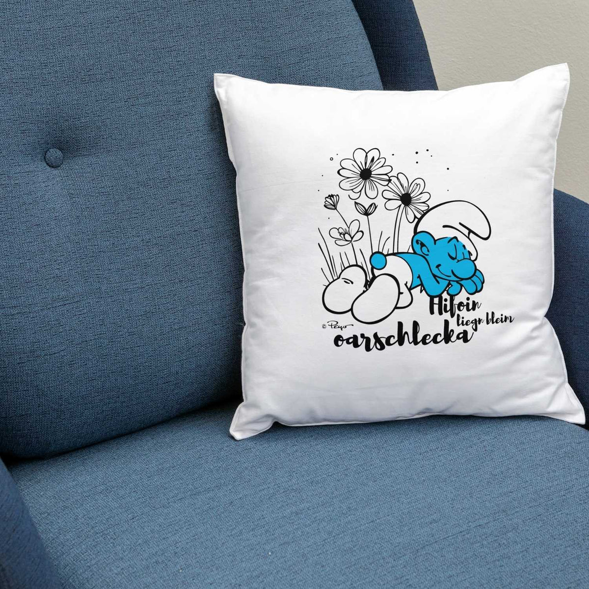Weißes ®Die Schlümpfe "Hifoin, liegn bleim, oarschlecka" Kuschelkissen mit schlafendem Schlumpf und Blumenmotiv auf blauem Sessel.
