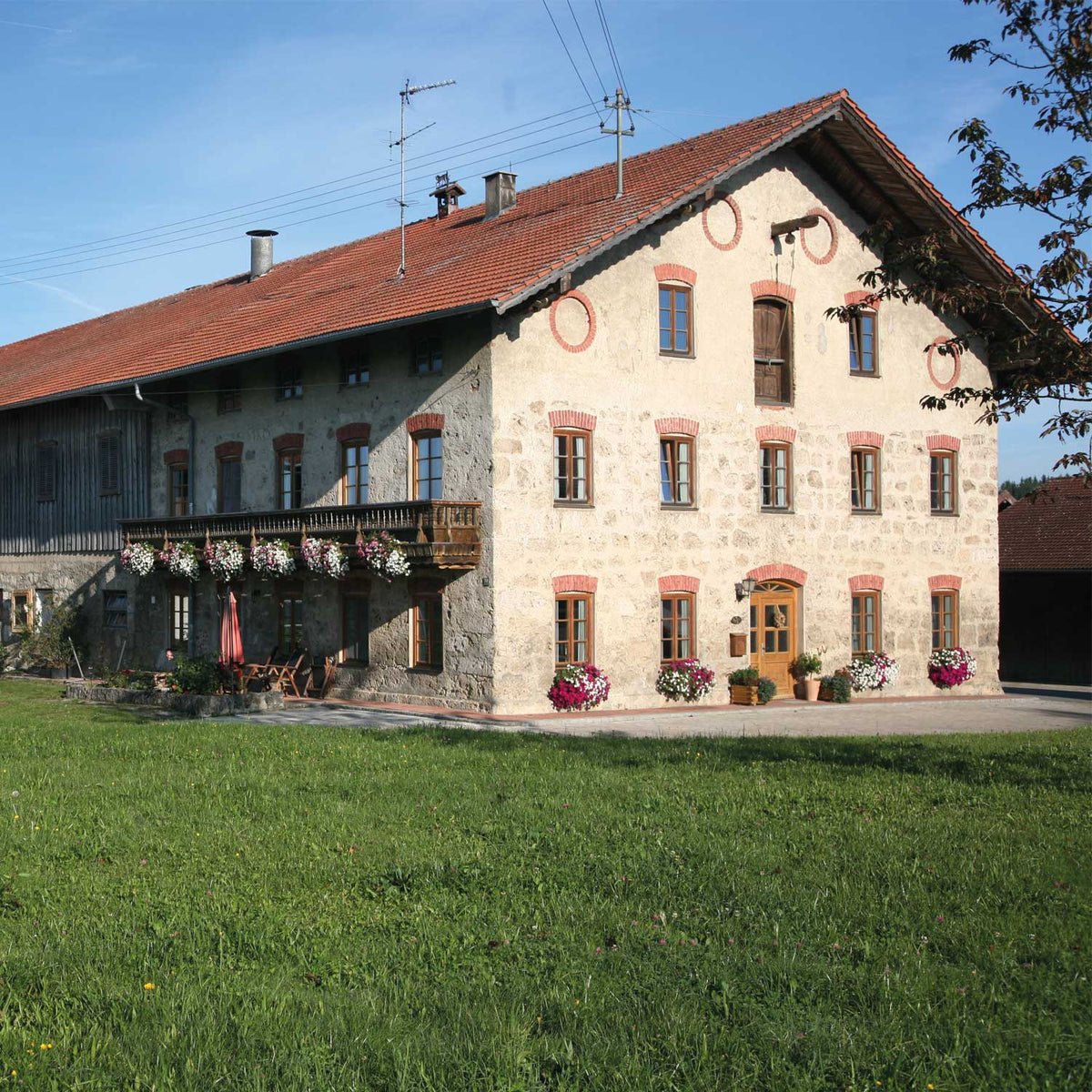 Traditionelles bayerisches Bauernhaus mit steinerner Fassade, Holzbalkon, roten Ziegeldach und blühenden Geranien.