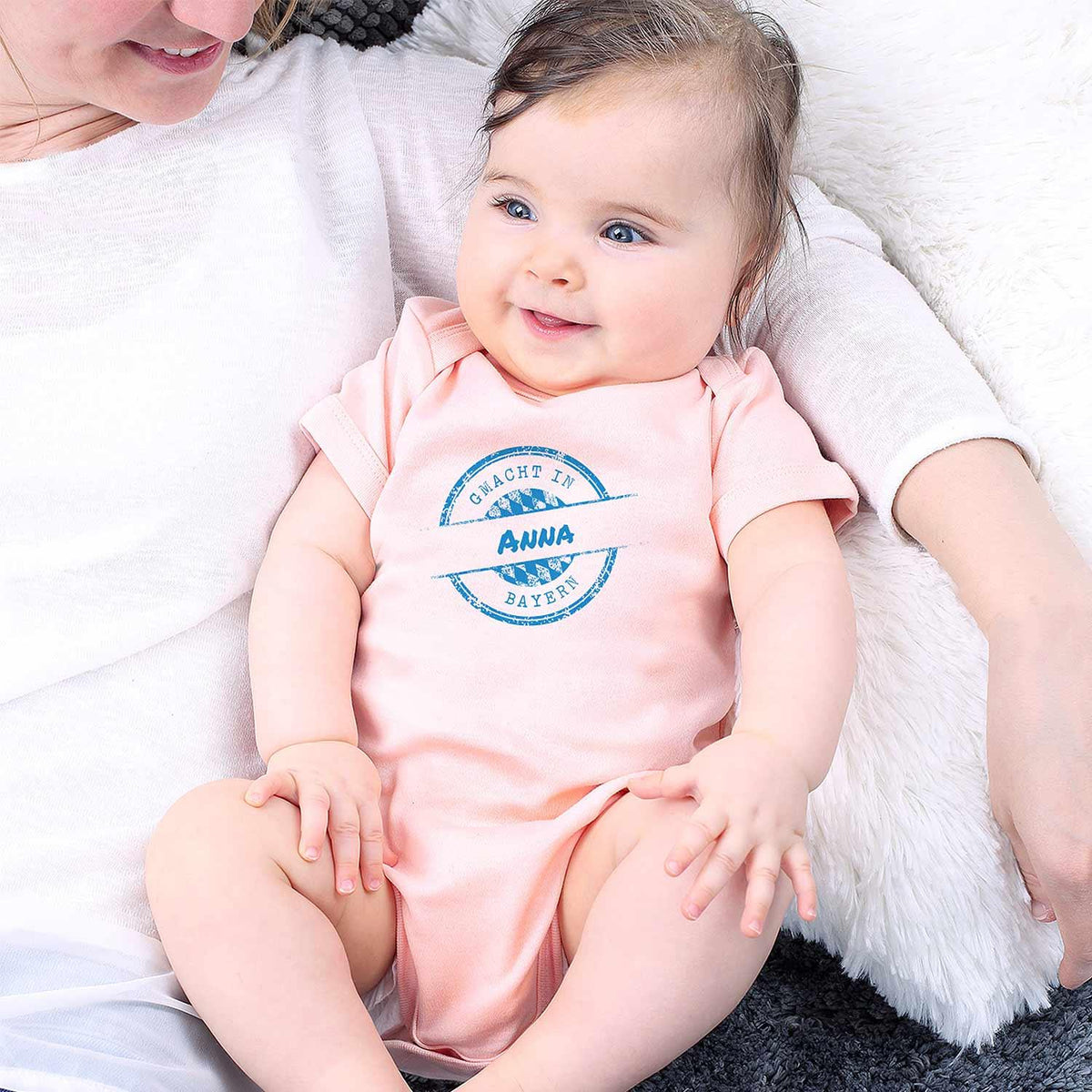 Glückliches Baby mit rosa Body g'macht in Bayern und personalisiertem Namen Anna, gehalten von einer Mutter.
