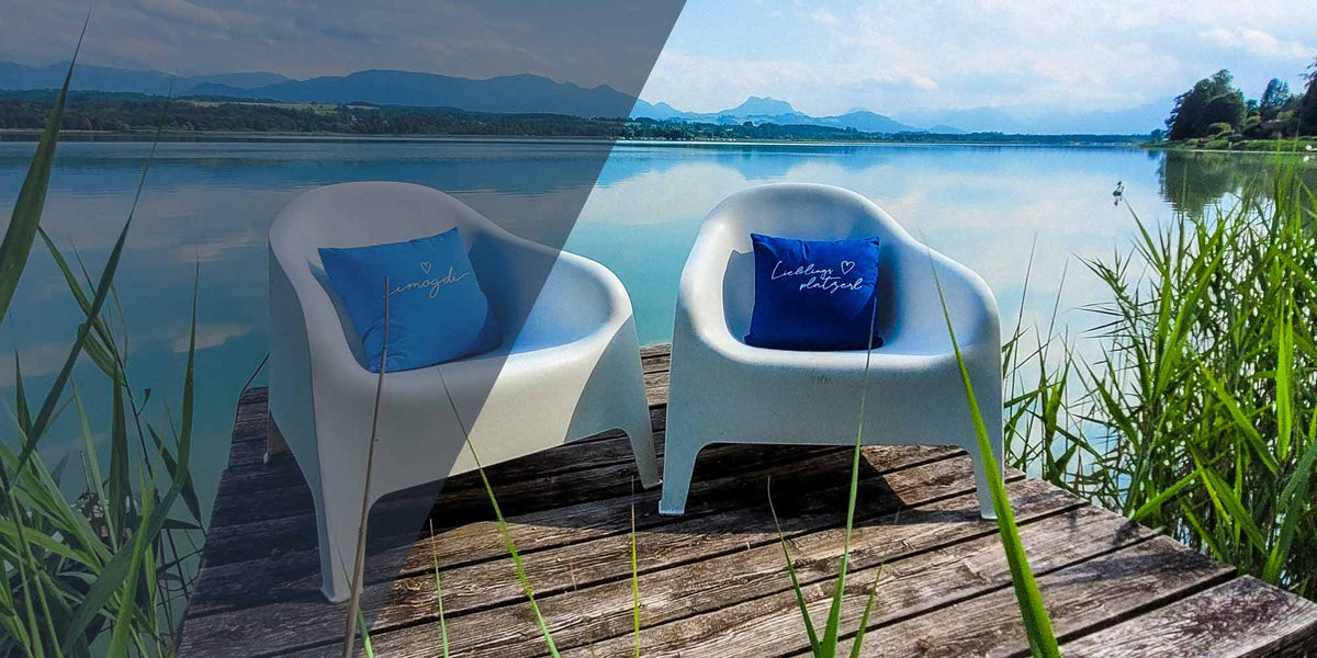 Zwei moderne Lounge-Sessel mit blauen Kissen auf einem Holzsteg am See mit Bergpanorama und Schilf.