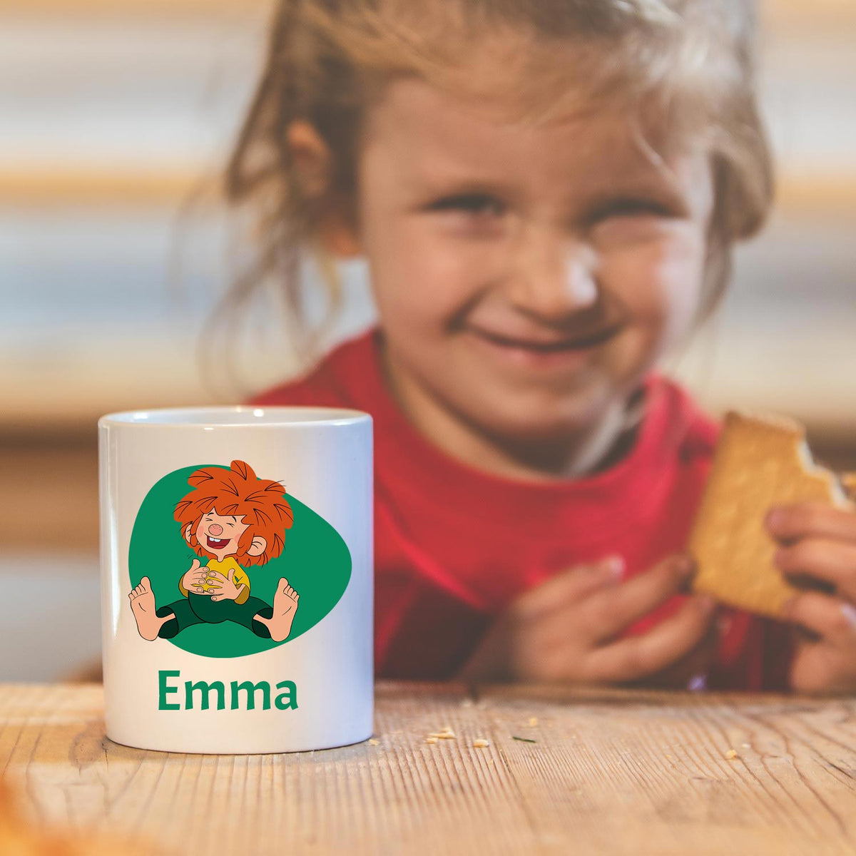 Weiße Pumuckl Tasse mit Wunschname Emma, sitzendem Pumuckl-Motiv und lachendem Kind im Hintergrund.