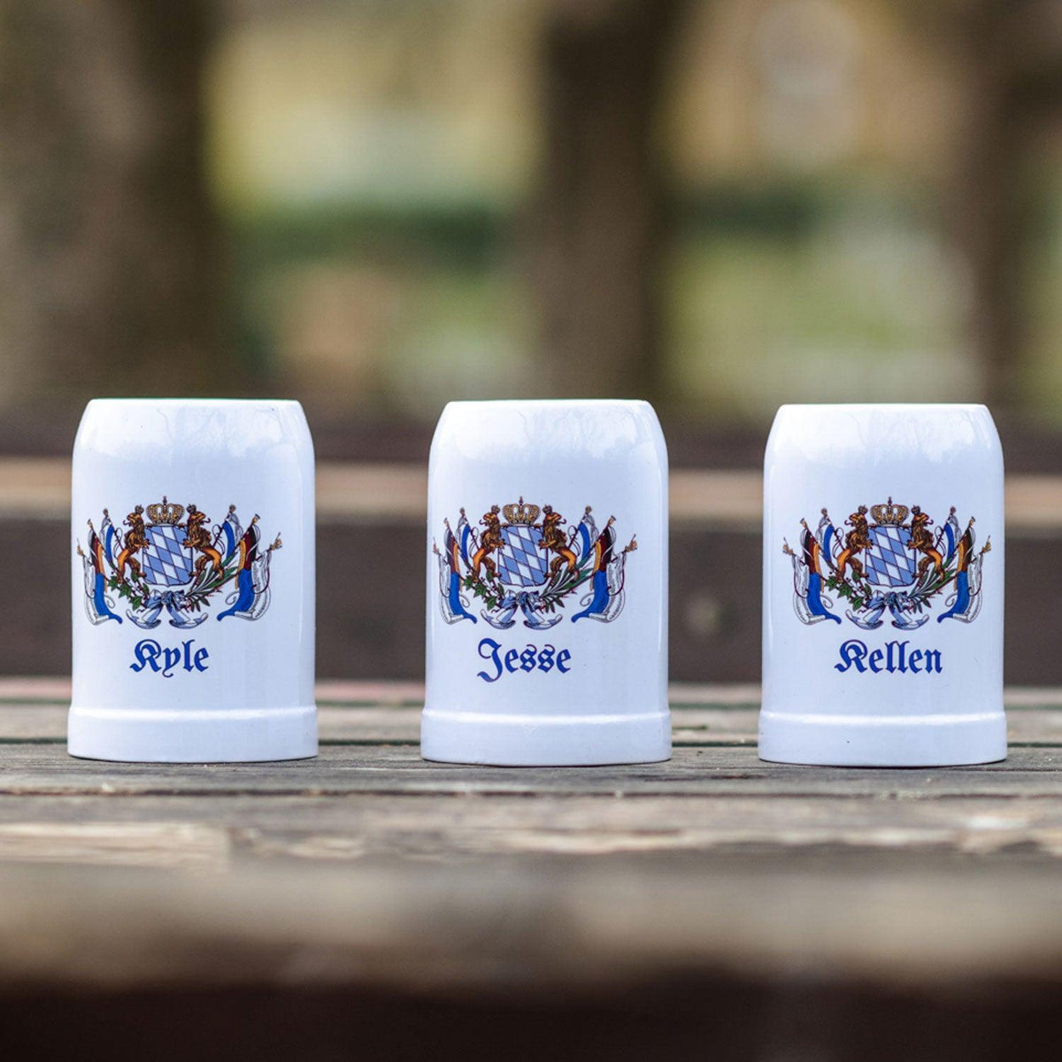Drei weiße Keramik-Bierseidel mit individuellen Namen und blau-weißem Bayernwappen auf Holztisch.