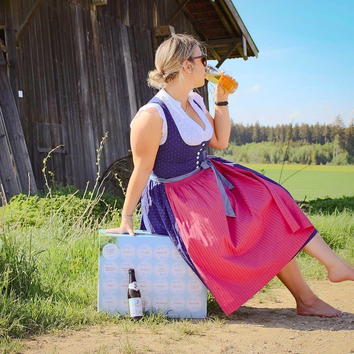 Frau im Dirndl sitzt auf der Bier-Box „Bayerischer Biergarten“ und trinkt Bier vor einer Holzhütte