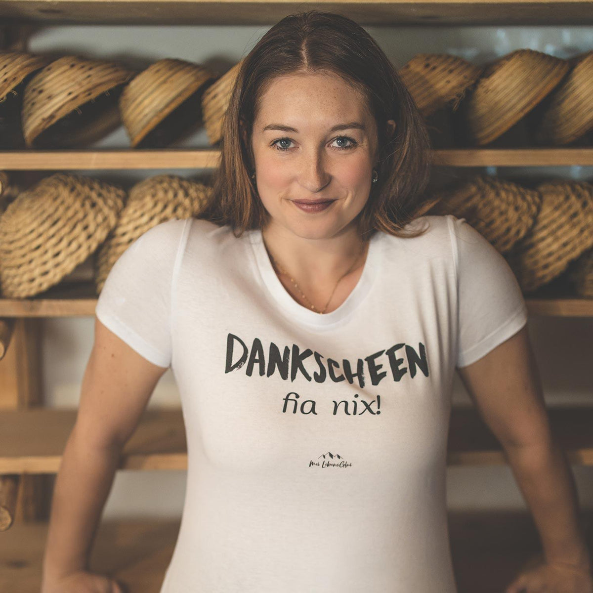 Lachende Frau trägt weißes Damen T-Shirt Dankscheen - fia nix! mit schwarzem Aufdruck vor Holzregal.