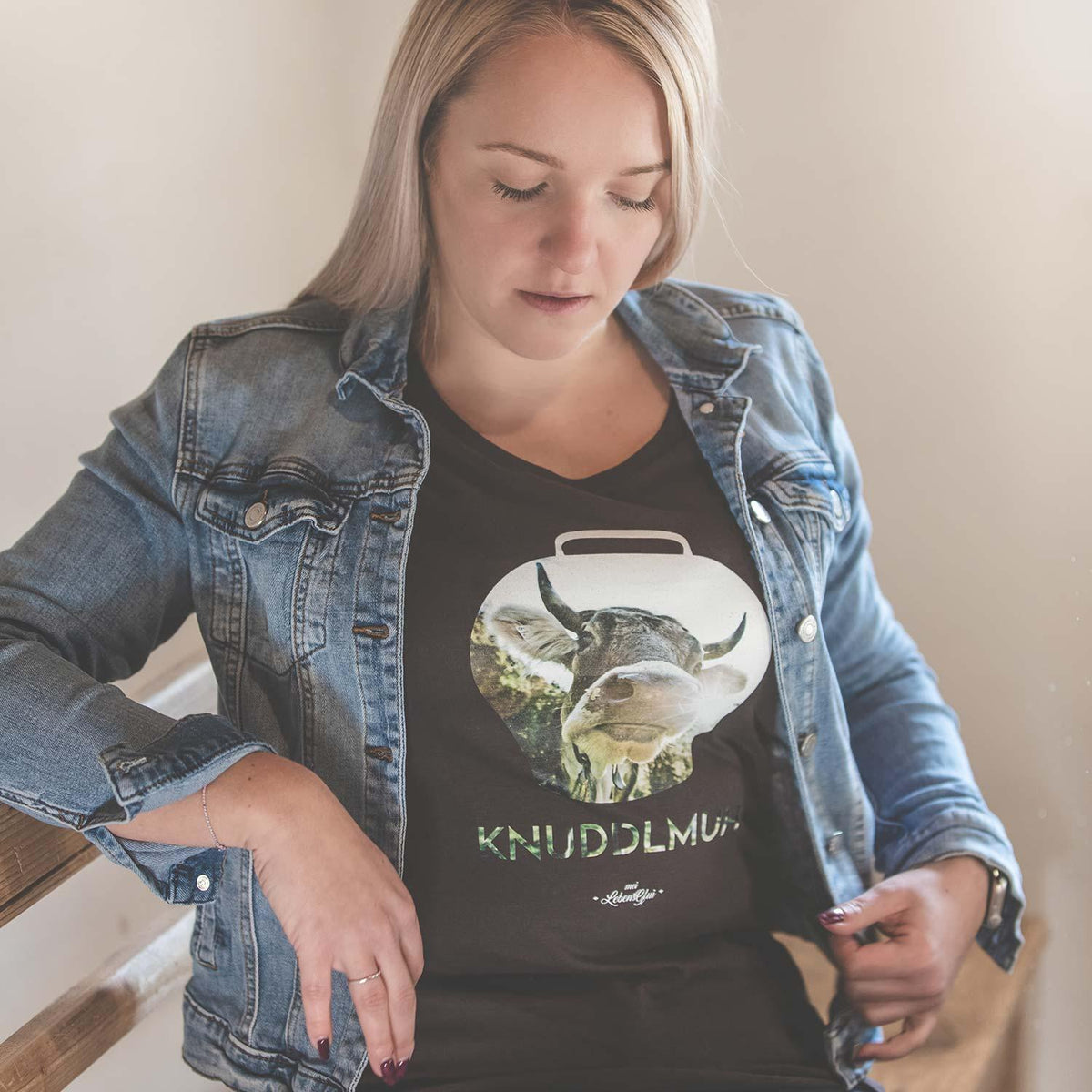 Blondes Modell trägt schwarzes Damen T-Shirt Knuddlmuh mit Kuh-Motiv in Jeansjacke, lässig angelehnt.