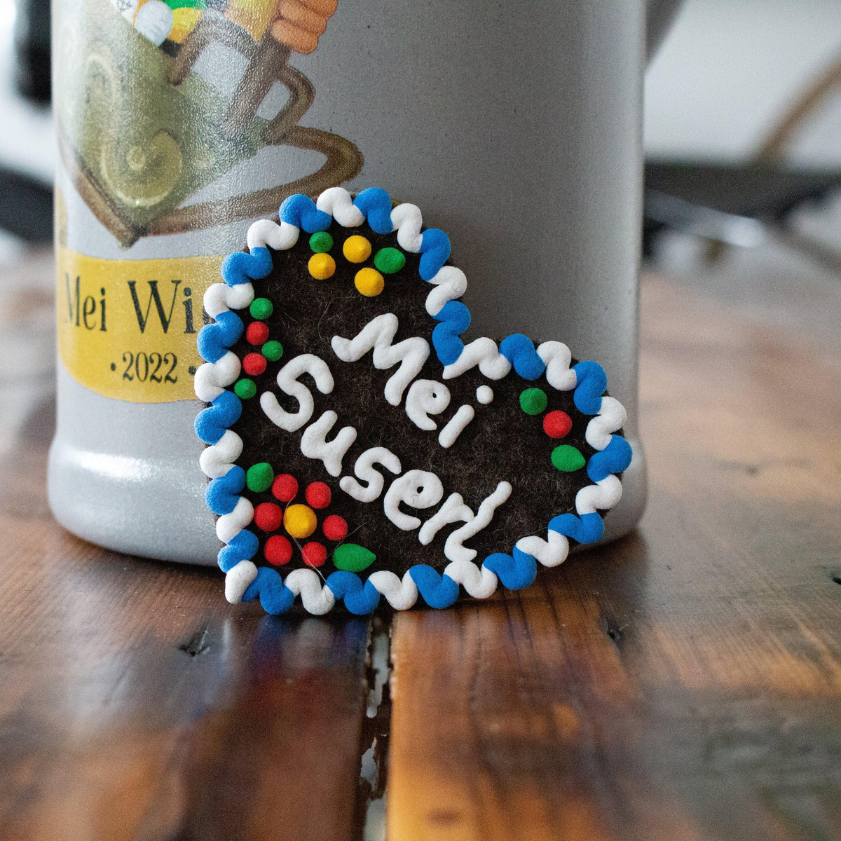 Filzherz-Anstecker Bayern mit Aufschrift „Mei Suserl“, weiß-blauem Rand und bunten Punkten auf einem Holztisch vor einem Bierkrug