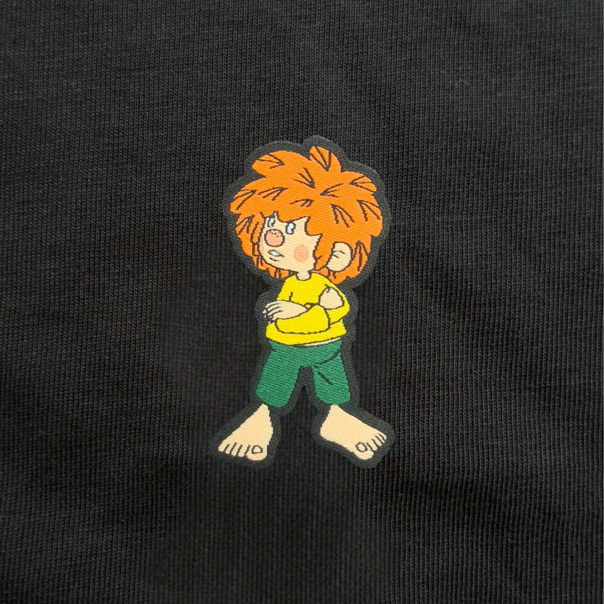 Nahaufnahme gewebtes Pumuckl-Label: grantiger Pumuckl mit verschränkten Armen auf schwarzem Shirt aus Bio-Baumwolle