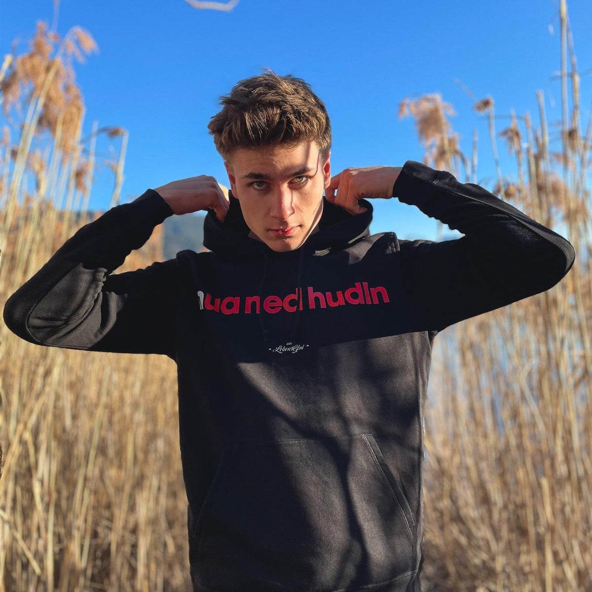 Junger Mann präsentiert coolen Herren Hoodie "Nua ned hudln" in Schwarz mit roter Schriftaufdruck outdoor.