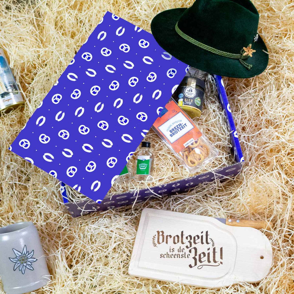 Geschenkbox mit bayerischen Spezialitäten, grünem Trachtenhut und Brotzeitbrett auf Heu.