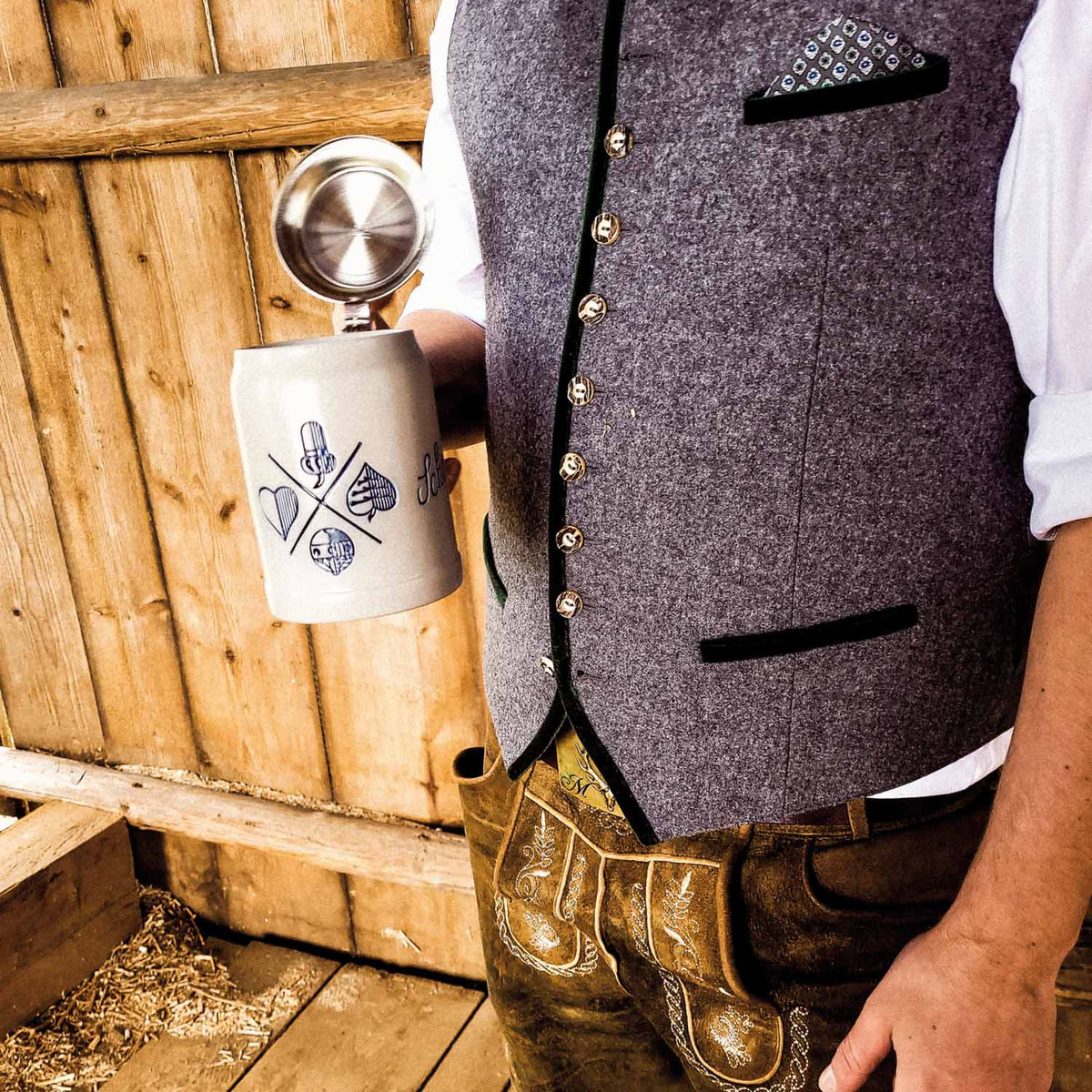 Person in traditioneller Tracht mit Lederhose, grauer Weste und weißem Hemd, hält einen verzierten Bierkrug mit Zinndeckel.