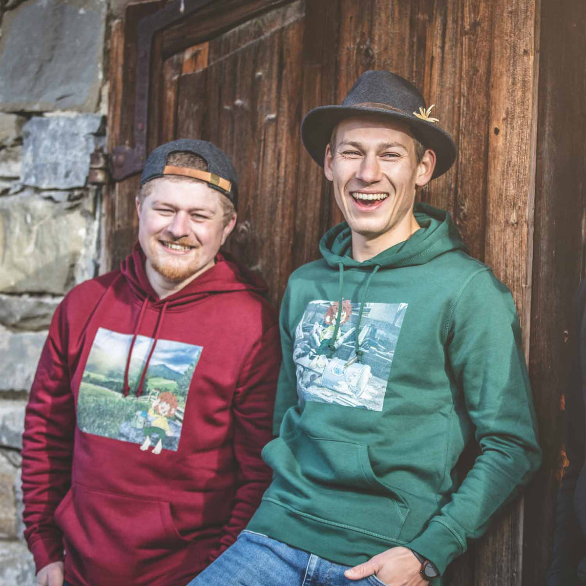 Zwei lächelnde Männer in bedruckten Hoodies lehnen an einer dunklen Holztür, tragen Hut und Kappe.