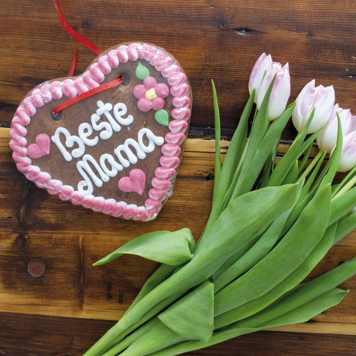Herzförmiges Lebkuchenherz mit Aufschrift "Beste Mama" und rosafarbenen Tulpen auf Holztisch.