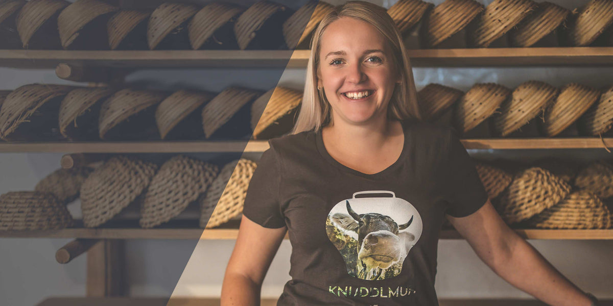 Freundliche junge Frau mit hellen Haaren und Kuh-T-Shirt vor Regalen mit geflochtenen Brotformen.
