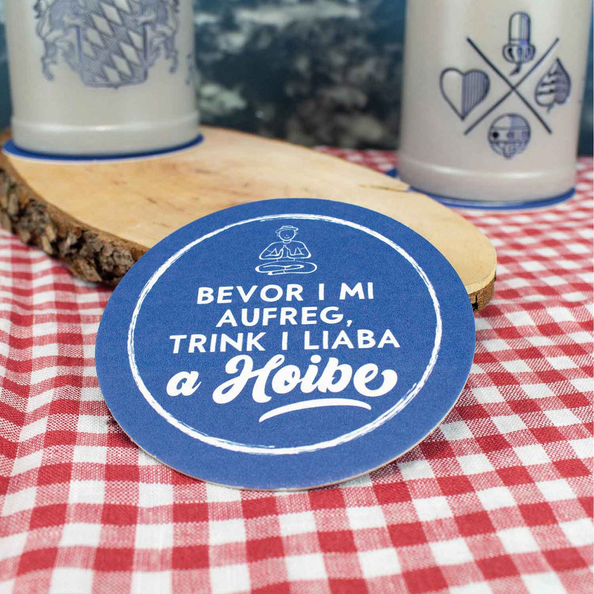Runder blauer Bierdeckel auf rot-weiß karierter Tischdecke mit originellem Spruch und Maßkrügen im Hintergrund.