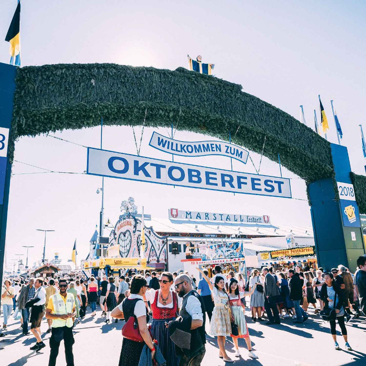 Großes grünes Oktoberfest Willkommensschild über belebter Festwiese mit vielen Besuchern, einige in Tracht.