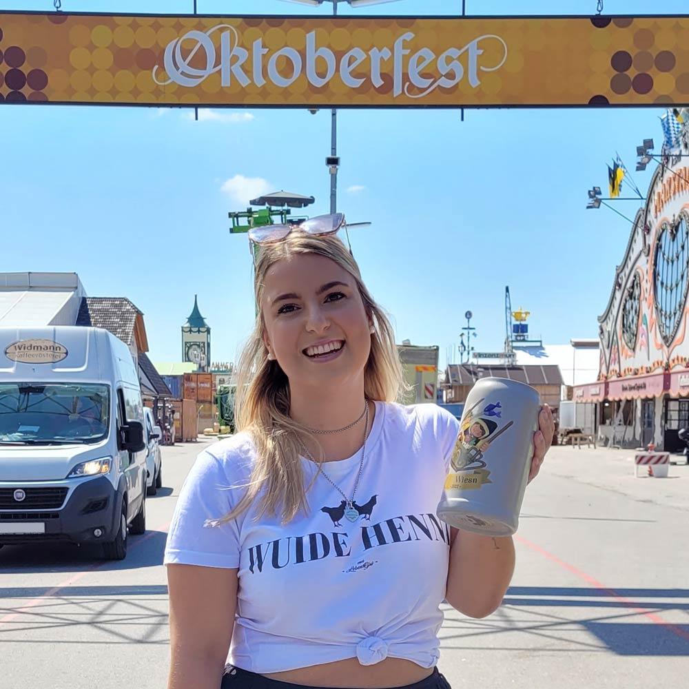 Junge Frau am Oktoberfest-Eingang hält stolz den grauen Mei Wiesn 2022 Sammlerkrug mit buntem Design.