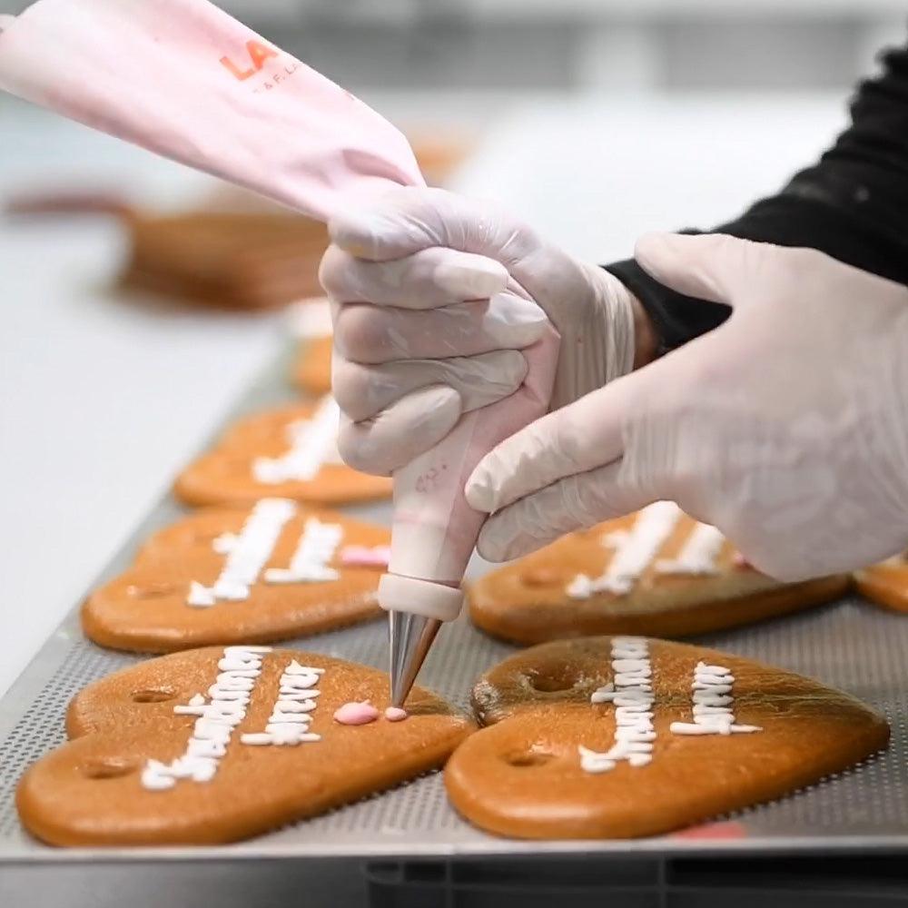 Nahaufnahme von Händen in weißen Handschuhen, die mit einem Spritzbeutel Lebkuchenherzen auf einem Backblech verzieren.