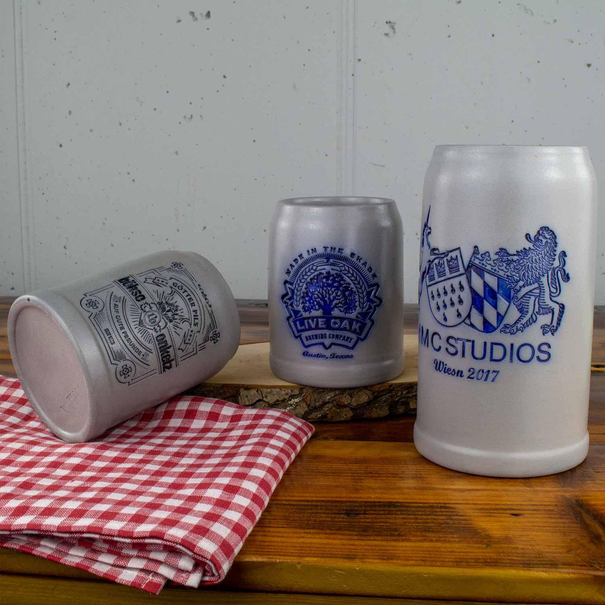 Drei graue und weiße Bierkrüge aus Keramik mit blauen Logos auf Holzplatte, daneben rot-weiß kariertes Tuch.