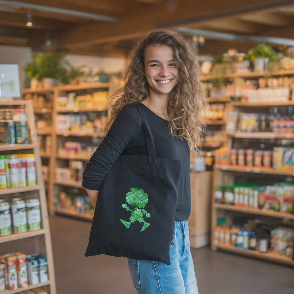 Lachende Frau mit schwarzer Stofftasche mit grünem Pumuckl Motiv in einem Naturkostladen.