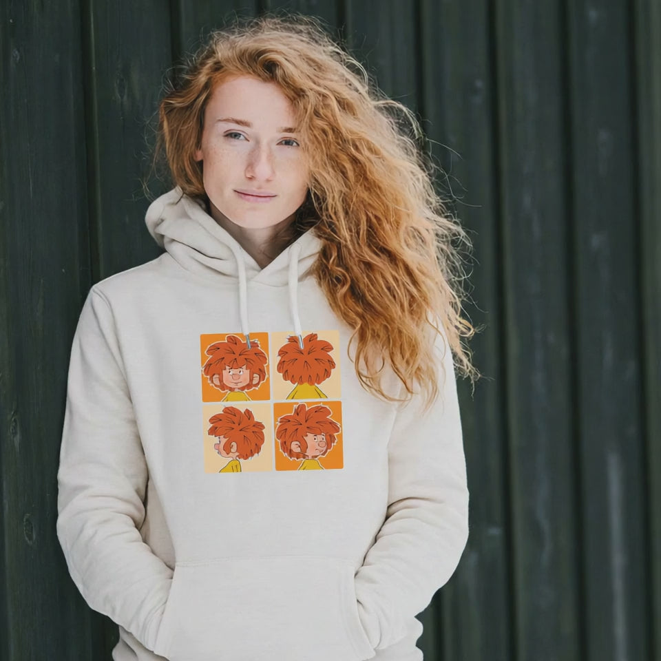 Frau mit roten Lockenhaaren trägt beigen Pumuckl Hoodie mit „Schnappschuss“-Motiv, lässig an Holzwand lehnend.