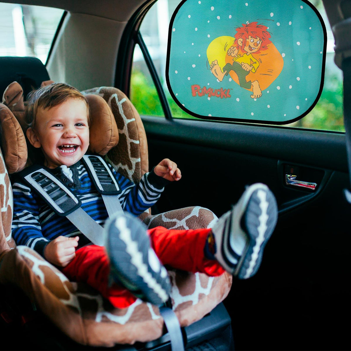 Kind lacht fröhlich im Kindersitz vor Pumuckl Auto Sonnenschutz mit buntem Motiv im Seitenfenster.