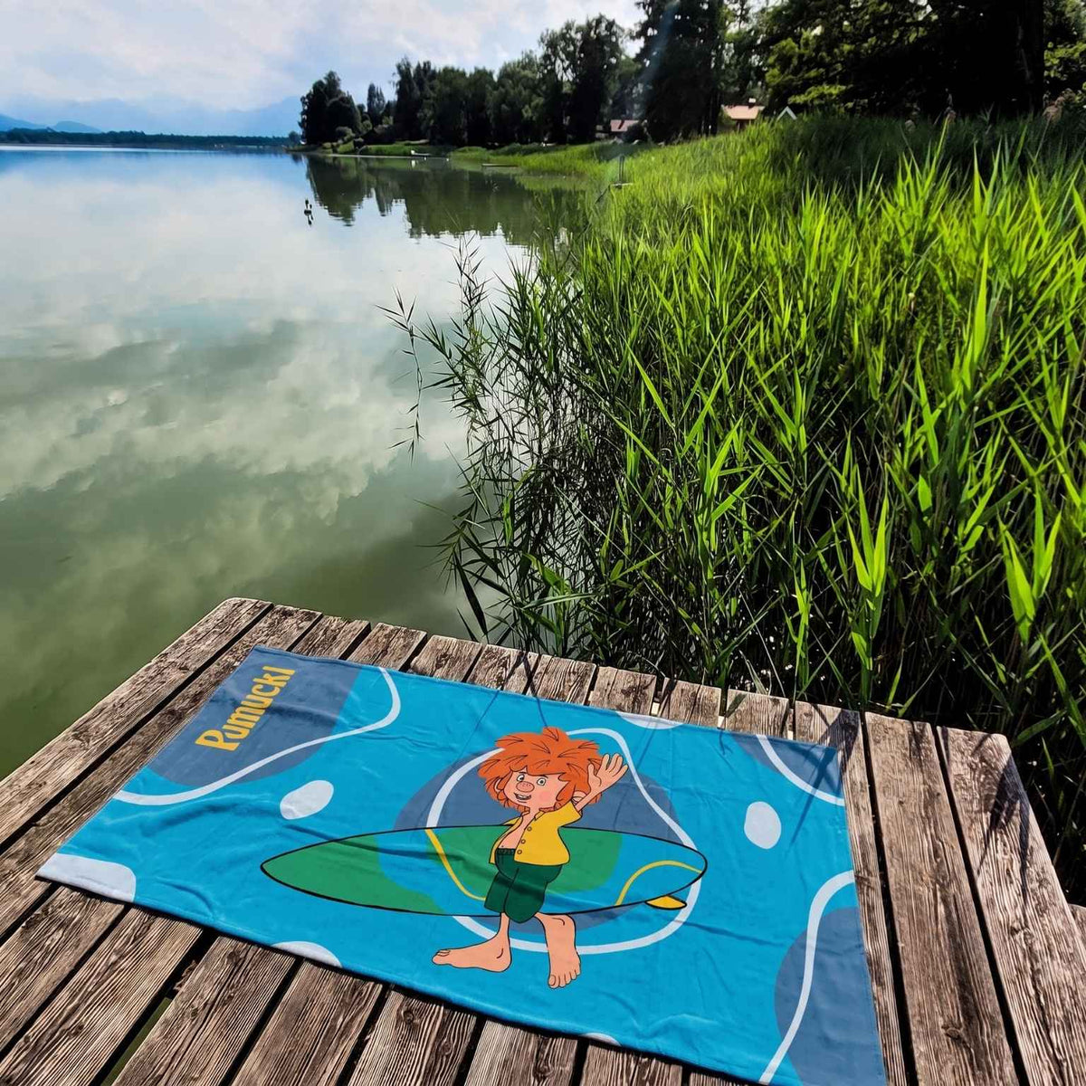 Pumuckl Handtuch XXL in Blau mit Surfbrett-Motiv auf einem Holzsteg am See – großes Badetuch 75 x 155 cm