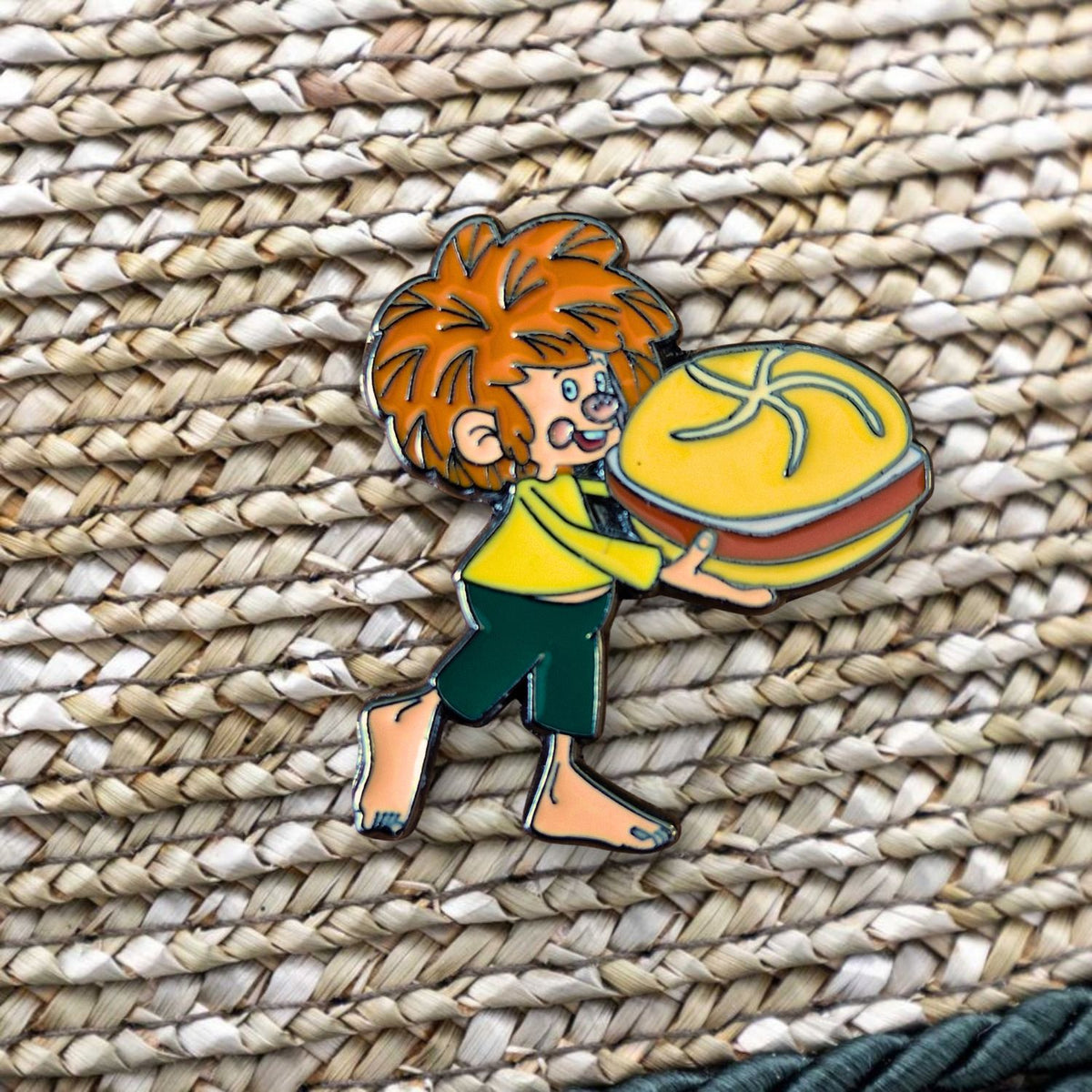 Pumuckl Pin Leberkassemmel – Emaille Anstecker: Pumuckl trägt eine Leberkassemmel im Arm, auf Strohgeflecht