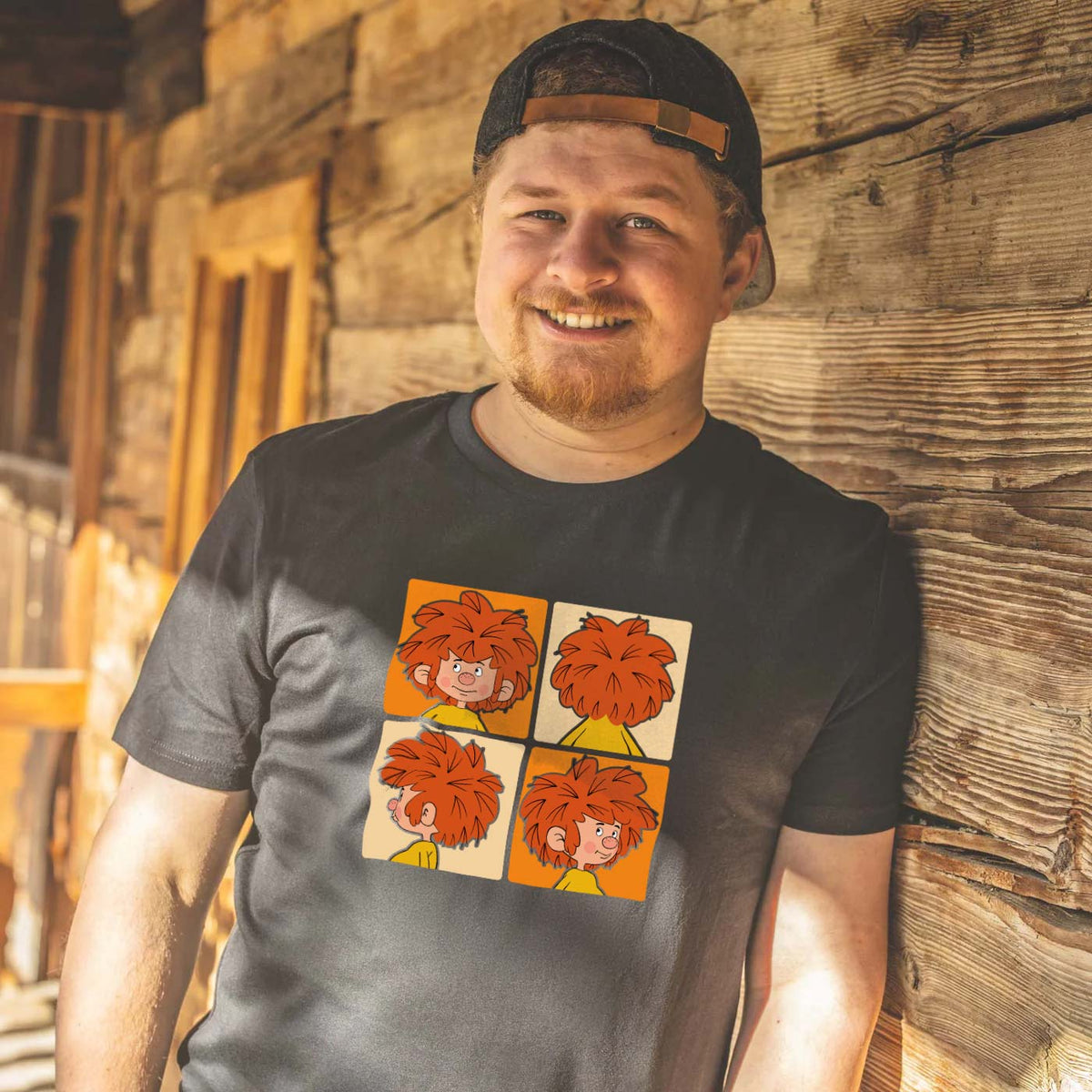 Lächelder Mann trägt schwarzes Pumuckl Herren T-Shirt Schnappschuss mit roten Kobold-Motiven vor Holzwand.