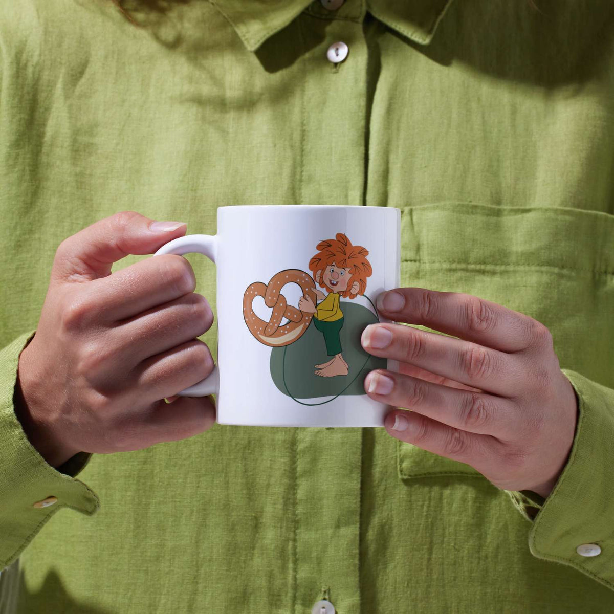 Pumuckl Tasse Breze aus Porzellan, in Händen gehalten, mit Pumuckl und einer Brezel.