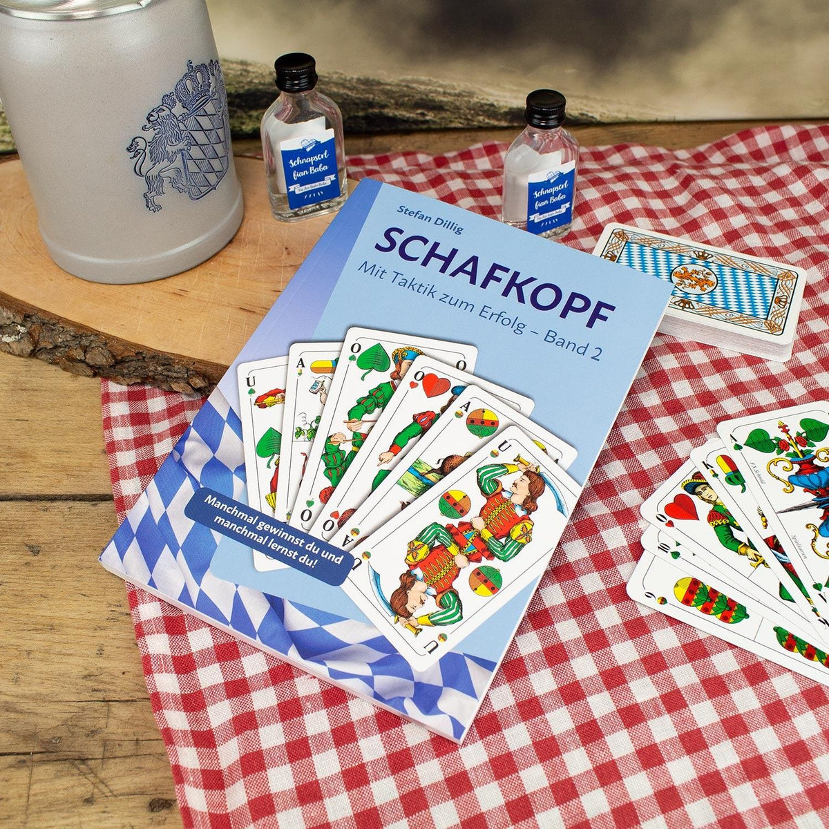 Schafkopf: Mit Taktik zum Erfolg - Band 2 Buch, Schafkopfkarten, Bierkrug und Schnapsfläschchen auf karierter Decke.