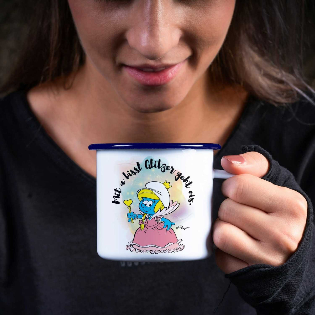 Junge Frau hält ®Die Schlümpfe Emailletasse "Glitzer" mit Schlumpfine-Motiv und Schriftzug in der Hand.
