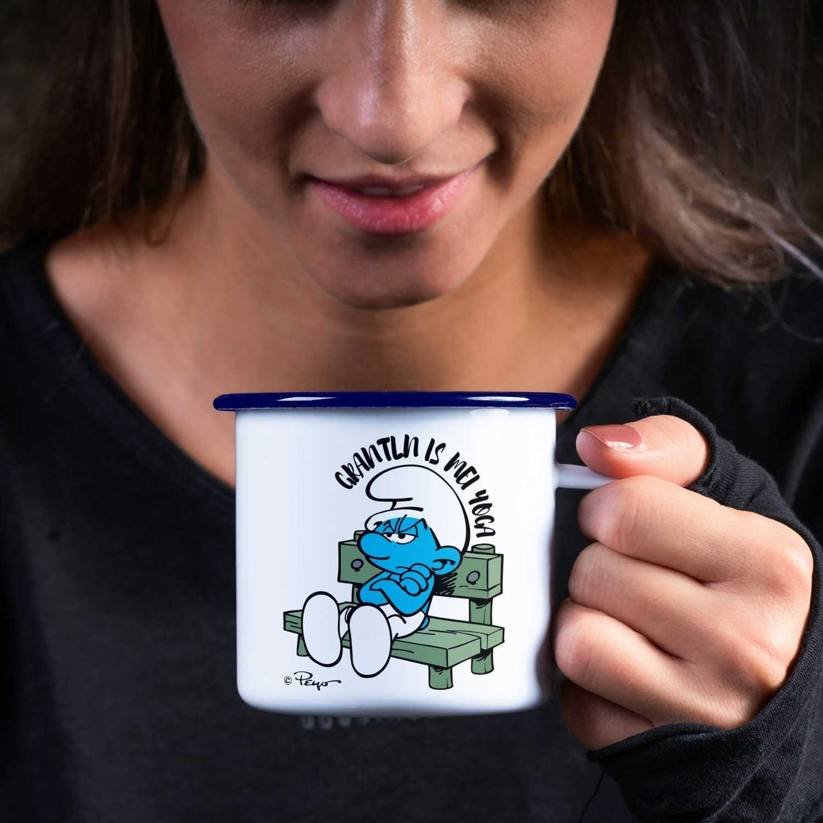 Dunkelhaarige Frau hält emaille Schlümpfe Tasse "Grantln is mei Yoga" mit mürrischem Schlumpf Motiv.