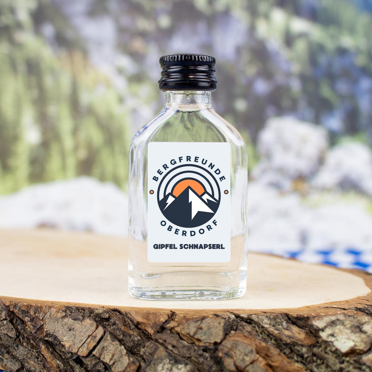 Kleine Schnapsflasche der Marke "BERGFREUNDE OBERDORF GIPFEL SCHNAPSERL" mit Bergsymbolik auf Baumscheibe.
